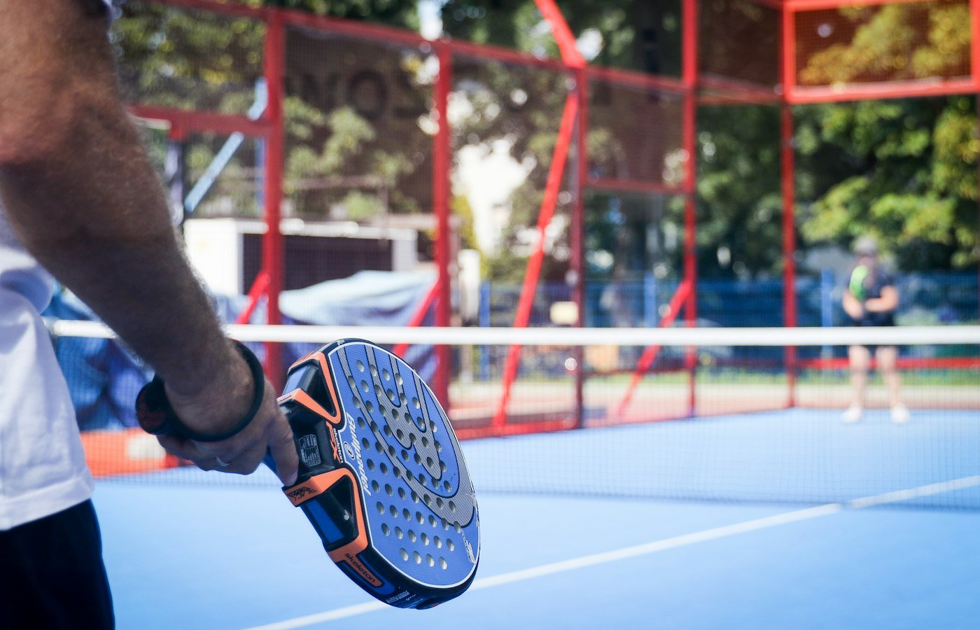 Main tenant une raquette de padel près du filet, joueur flou en arrière-plan sur un court bleu
