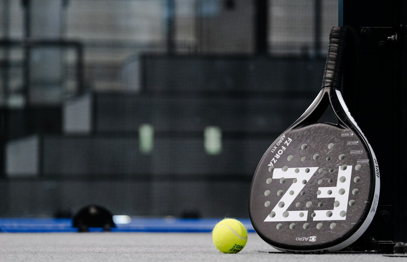 Raquette de padel noire FZ et balle jaune près d’un poteau sur une piste claire