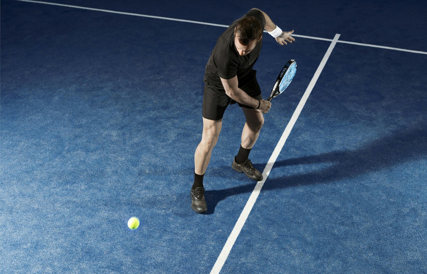 Joueur de padel en noir frappant une balle basse sur un court bleu, vu de dessus