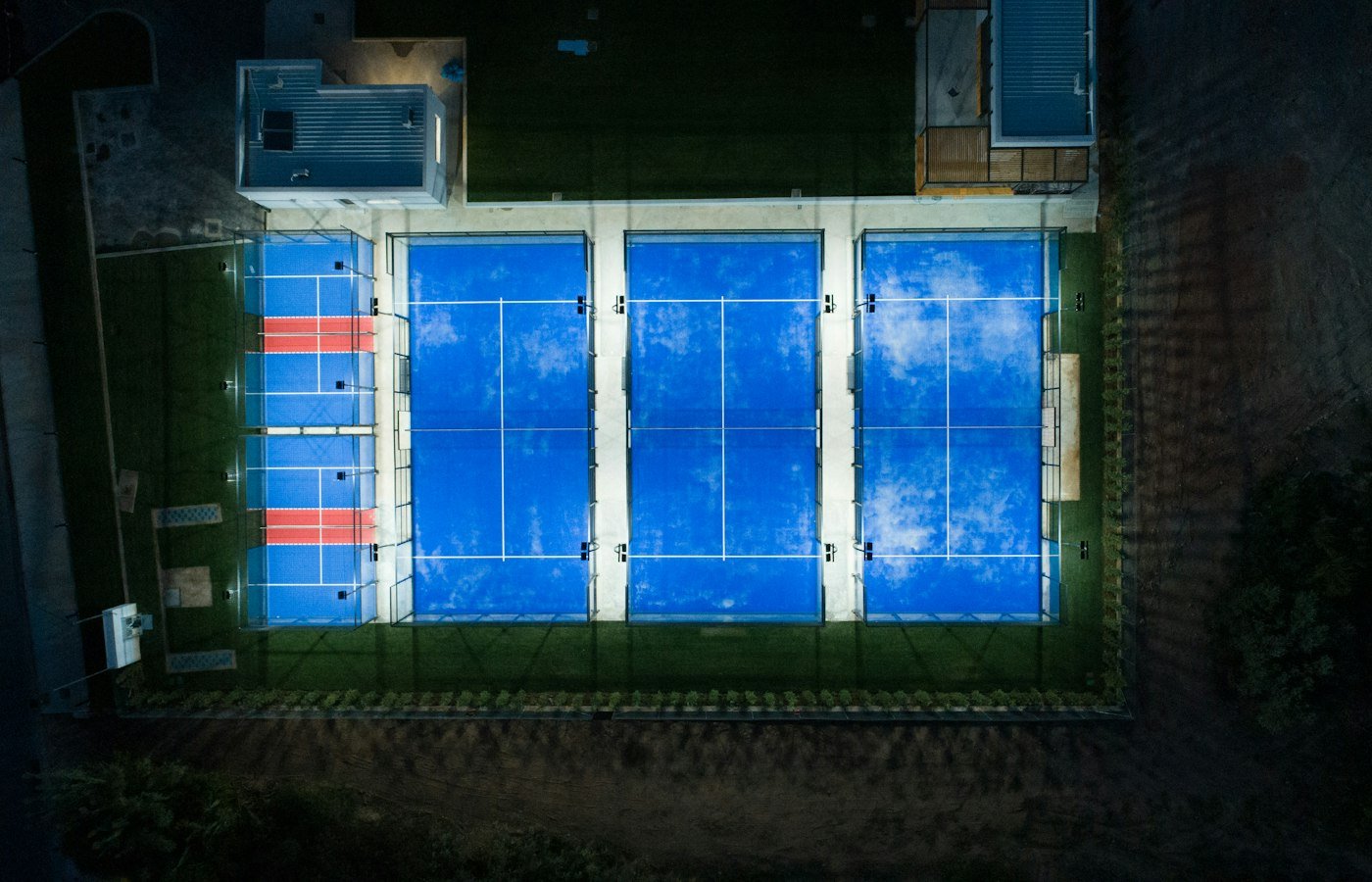 Vue aérienne de nuit de trois terrains de padel bleus éclairés, entourés de vitres
