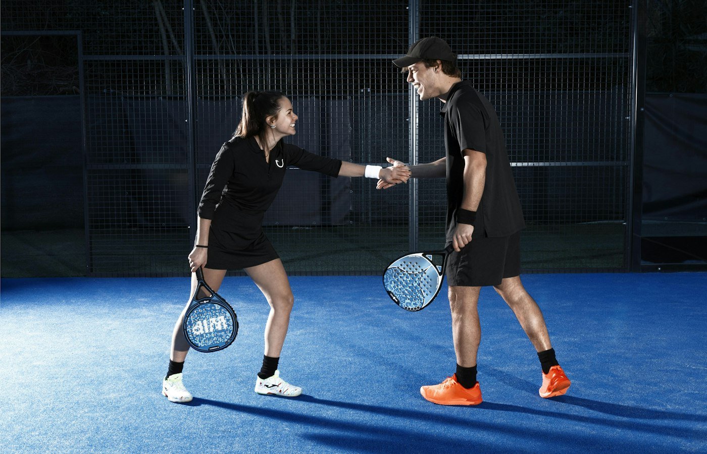 Homme et femme se saluant d’un check sur un court de padel bleu, raquettes à la main
