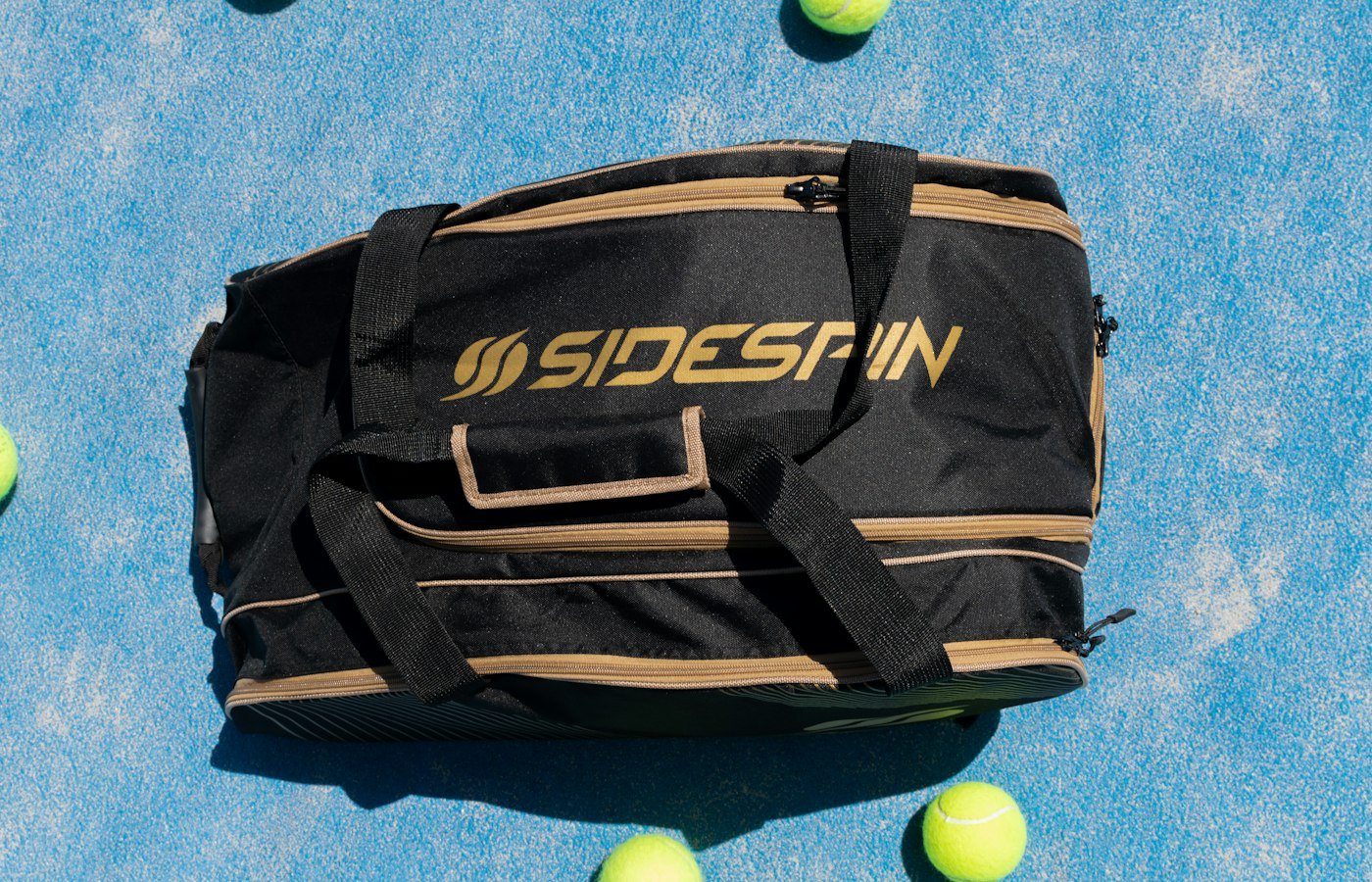 Sac de padel noir et doré posé sur une piste bleue, entouré de balles de padel