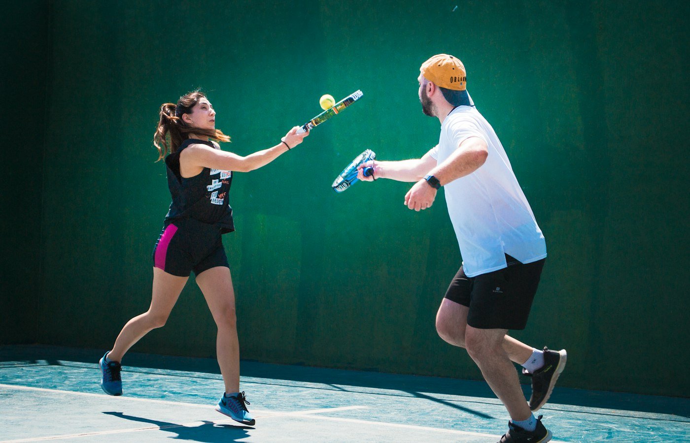Un homme et une femme échangent au padel devant un mur vert, raquettes pleines et balle en l’air