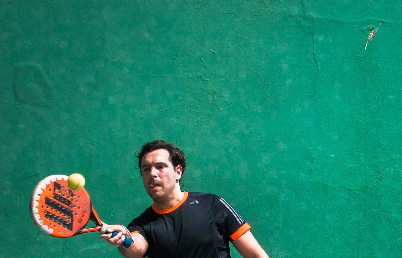 Joueur de padel en t-shirt noir qui frappe une balle avec une raquette orange devant un mur vert