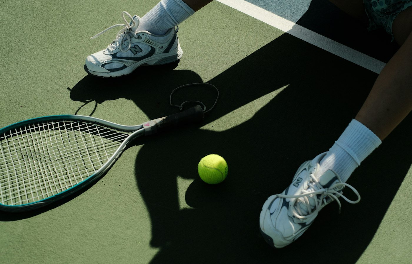 Pieds d’un joueur en baskets blanches sur un court vert, raquette et balle au sol