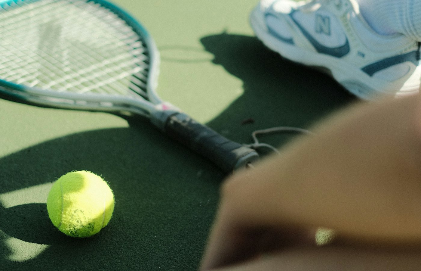 Raquette posée sur un court vert près d’une balle jaune, pieds d’un joueur en baskets