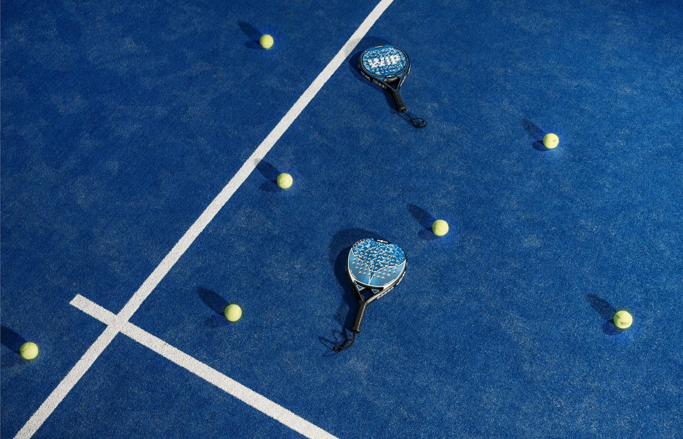 Deux raquettes de padel bleues et des balles jaunes posées sur le gazon synthétique d’une piste